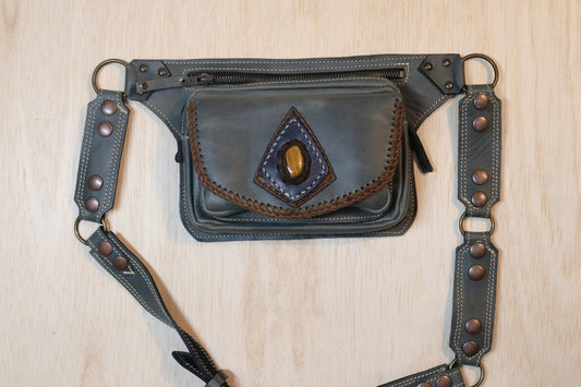 A dark gray leather belt bag with a front flap pocket featuring a triangular brown leather patch and a central amber-colored stone. The bag has a top zipper, metal rings, and an adjustable leather strap with copper-toned rivets, laid flat on a light wood background.