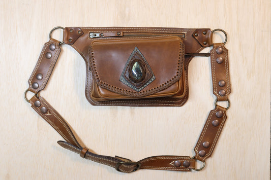 A brown leather fanny pack with a decorative concho-style emblem on the front flap, a top zipper, and an adjustable leather strap with metal rivets, laid flat on a light wood background.